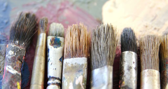 A collection of used paintbrushes with dried paint on them rests against a colorful, paint-splattered background. These tools reflect the messy and creative process inherent in painting and artistry.