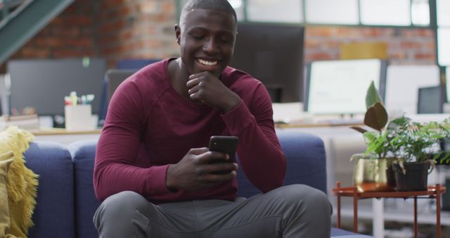 This image features a young man sitting in a modern office lounge area, smiling while reading something on his smartphone. This engaging and positive scene can be used for promoting work-life balance, illustrating modern communication methods, or adding a human touch to business-related content. It is suitable for marketing, workplace culture features, and technology-related messages.