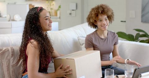 Two african american women unpacking moving box on couch in bright cozy living room