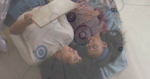 Smiling mother and daughter lying together holding tablet, cozy family bonding at home