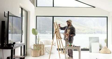 Indian artist painting on easel in bright lakeside modern studio with panoramic window