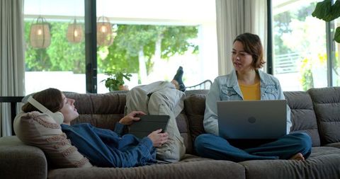 Diverse young women relaxing on sofa with laptop and tablet collaborating from home for remote study