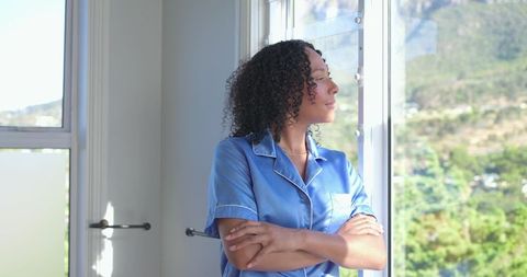 African woman in blue pajamas gazing through sunlit window with blinds, serene morning