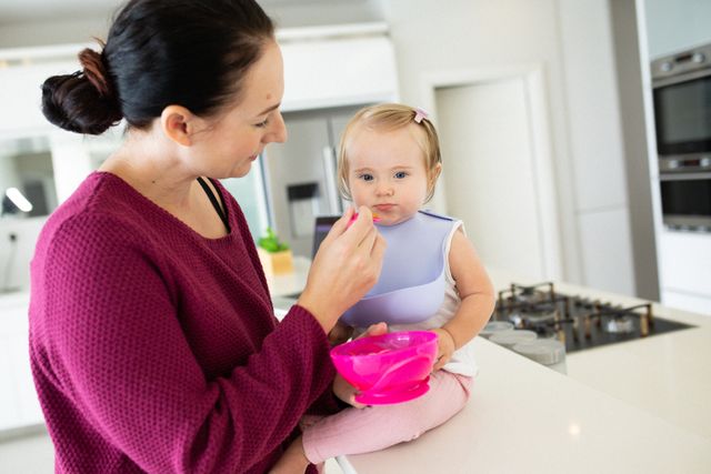 Happy caucasian mother feeding baby daughter sitting on counter top in kitchen. at home in isolation during quarantine lockdown.