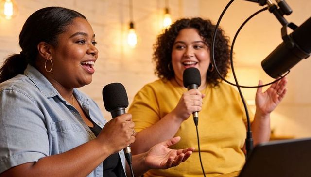 Two co-hosts recording lively podcast in cozy warm-lit studio with boom microphone