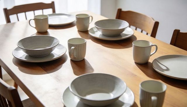 Sunlit wood dining table showcasing ceramic dinnerware and ribbed mugs, minimalist