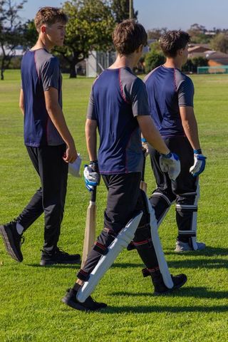 Teen cricket players walking across sunlit field carrying bats wearing navy team uniforms