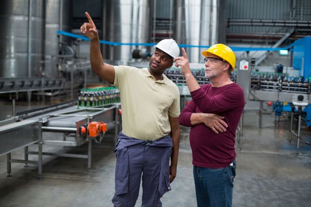 Two factory workers in safety helmets are discussing and pointing upwards in a beverage production plant. This image can be used to illustrate teamwork, industrial processes, workplace safety, and professional collaboration in manufacturing environments.