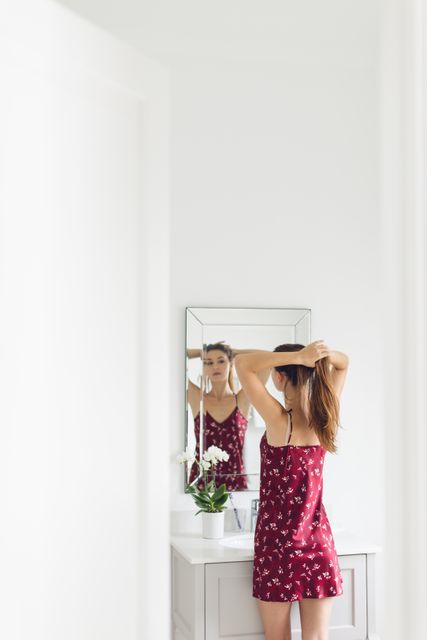 Woman stands in front of bathroom mirror, tying her hair. Ideal for themes related to morning routines, self-care, elegance, and home life. Excellent for blogs or articles focused on personal grooming, lifestyle, or interior design.