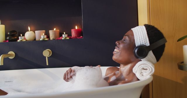 Woman enjoying bath time in a luxurious tub with foam bubbles. She wears headphones and smiles, savoring the moment. Background features various lit candles adding to the tranquility. Ideal for illustrating concepts of self-care, relaxation, wellness, spa atmosphere, luxury, and personal time.
