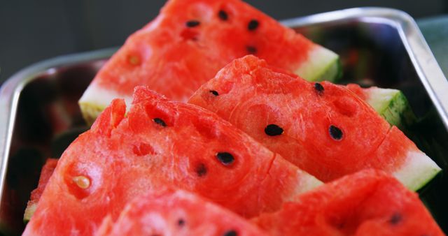 Slices of ripe watermelon are arranged in a metal tray, showcasing the vibrant red flesh and black seeds. Watermelon is a refreshing and popular fruit enjoyed for its juicy sweetness and hydrating properties.