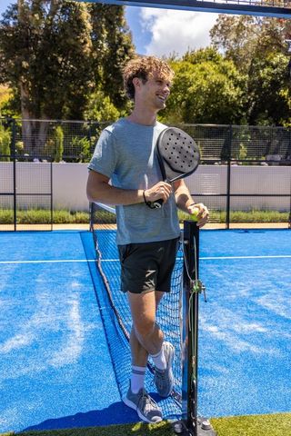 Smiling young man leaning on net holding padel racket and ball on vibrant blue court