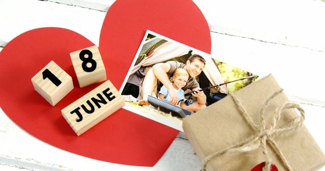 Symbolizing a Father's Day celebration, this composition features a wrapped gift in brown paper tied with twine, a large cut-out red heart, and wooden blocks displaying June 18. The addition of a family photo accentuates themes of love and bonding. Ideal for promotional materials, greeting card imagery, and holiday marketing campaigns.
