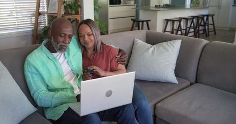 Mature couple sharing laptop on sofa, using smartphone for online banking and shopping