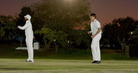 Two cricketers appealing on dusk floodlit pitch wearing white uniforms and bucket hat