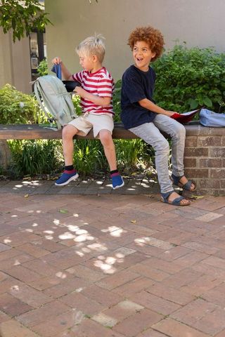 Two diverse boys sitting on bench in school courtyard laughing with backpack and folder