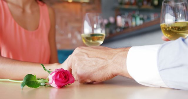 A Caucasian couple enjoys a romantic moment over glasses of wine, with a single rose symbolizing affection on the table, with copy space. Their hands touching gently convey a sense of intimacy and connection.