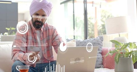 Man wearing pink turban analyzing laptop data while holding smartphone with hud overlays