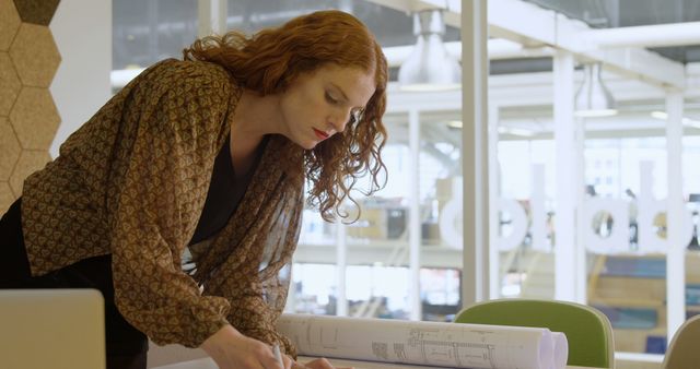 Focused young Caucasian woman reviews architectural plans in an office. She's immersed in her work, exemplifying dedication in a professional setting.