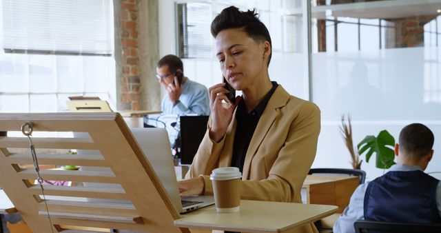 Businesswoman multitasking on phone call and laptop, coffee cup by her side, in a modern workspace with colleagues in the background. Perfect for corporate, startup and productivity themes.