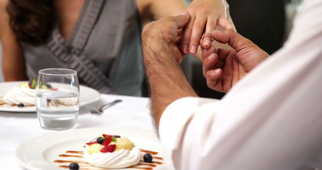 Man proposing marriage by placing a ring on woman's finger at a romantic dinner, captured in close-up. Delicious desserts and glasses of water set on white-clothed table enhancing the romantic setting. Ideal for illustrating concepts of love, marriage proposals, engagements, and romantic dining experiences.