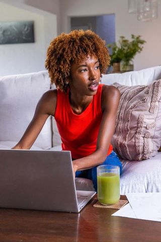 African american woman working remotely on laptop while leaning on couch with green juice