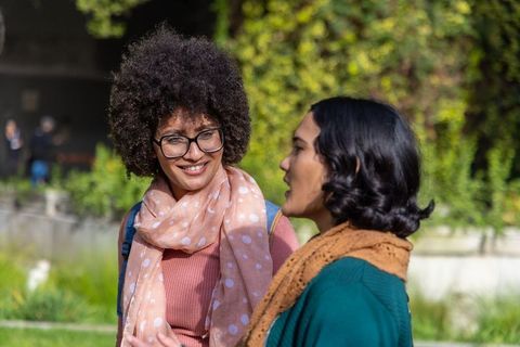 Two young women chatting and walking in sunlit park wearing scarves and glasses