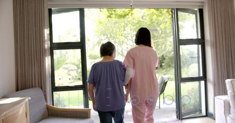 Caregiver Assisting Elderly Woman Walking Toward Sunlit Garden Through Patio Glass Doors