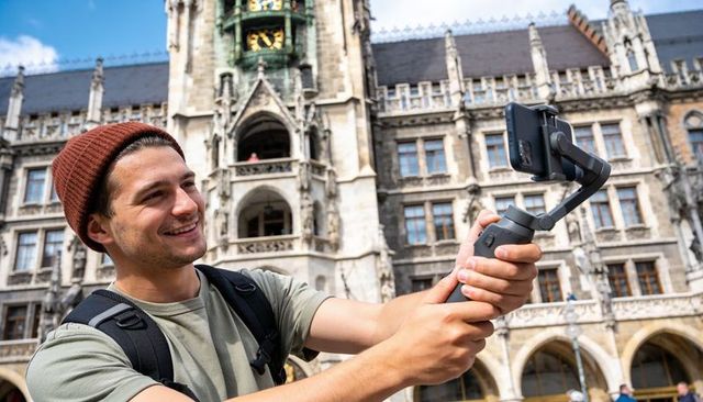 Smiling male traveler recording vlog with smartphone gimbal in historic gothic city square