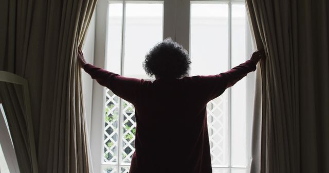Person standing in sunlit room, opening curtains to let in natural light. Useful for themes related to morning routines, relaxation, home interior, and starting the day on a positive note.