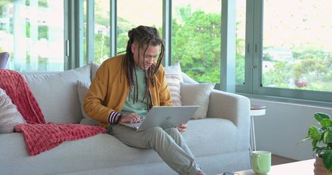 Man with dreadlocks working on laptop in bright living room for remote work productivity