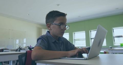 Young student typing on laptop in classroom wearing glasses focusing on digital learning