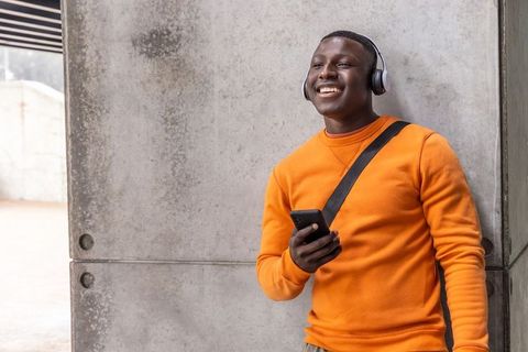 Smiling african american man listening to music with headphones leaning on concrete wall