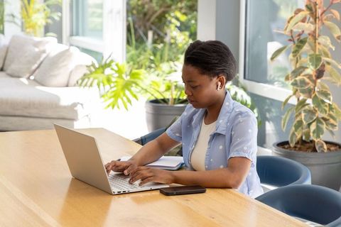 Teenage female working on laptop with phone and notebook at bright indoor plant workspace