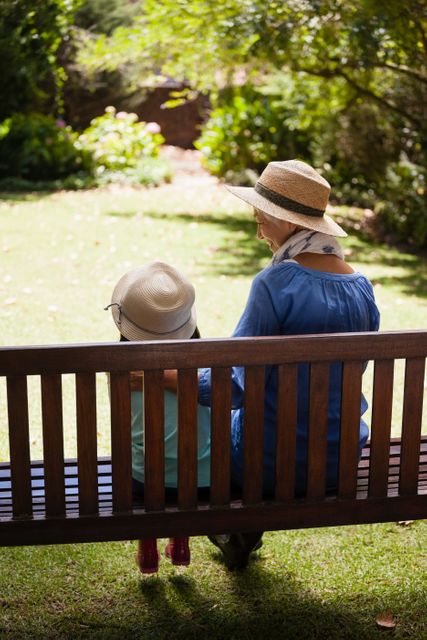 Rear view of granddaughter and grandmother wearing hats sitting on wooden bench at backyard