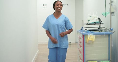 Smiling female nurse walking hospital corridor in blue scrubs passing crash cart