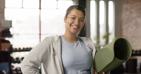 Young woman smiling holding green yoga mat in urban gym loft wearing gray hoodie