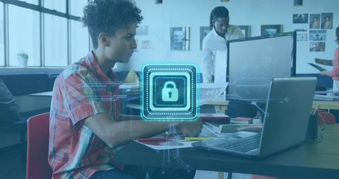 Young man working on laptop in coworking space with holographic cybersecurity lock overlay
