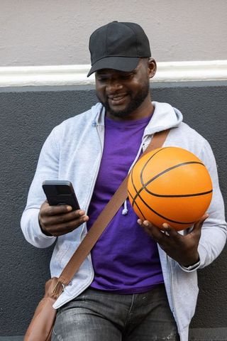 African man holding basketball and checking smartphone while leaning on urban wall