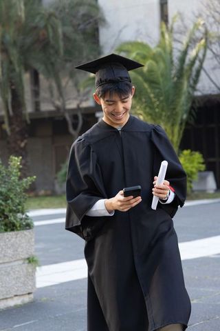 Asian male graduate smiling while checking phone and holding diploma on university campus