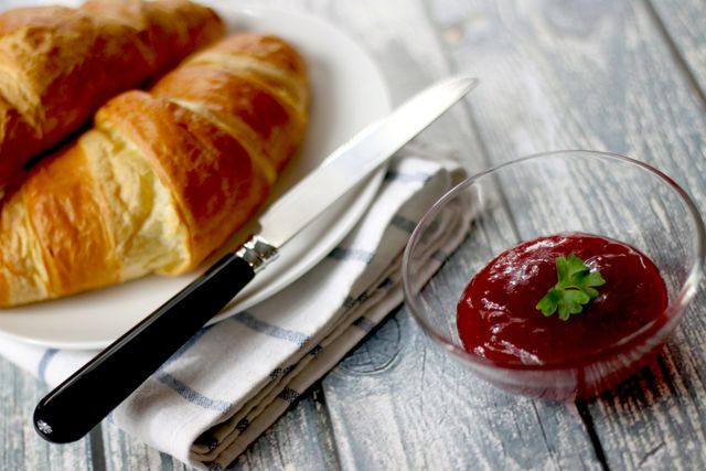 Fresh buttery croissants on white plate, alongside fruit jam in small glass bowl with garnish, and knife on plaid napkin, presented on rustic wooden table. Perfect for depicting cozy breakfast scenes, bakery promotions, and food recipes.