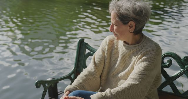 An elderly woman sits alone by a tranquil lake, basking in the sunlight and reflecting. Her thoughtful posture suggests a moment of introspection and calmness. Great for concepts of peaceful aging, enjoying nature, and solitude. This image is suitable for use in health and wellness articles, retirement community materials, and mindfulness content.