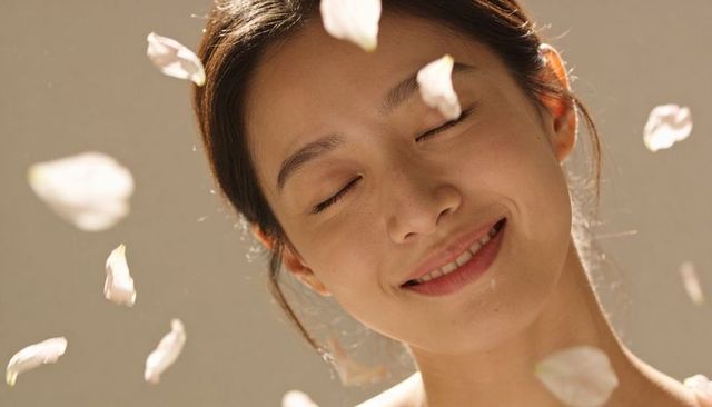 Serene asian woman smiling with eyes closed, falling blossom petals, warm studio light