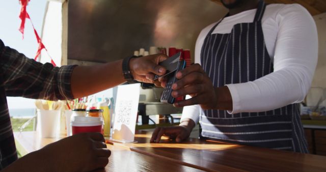 Customer making a contactless payment at a food truck with a bank card. This image showcases modern payment technology in the food service industry, with the employee wearing a stripe apron. Ideal for articles or advertisements about mobile dining, cashless transactions, and convenient payment methods in restaurants.