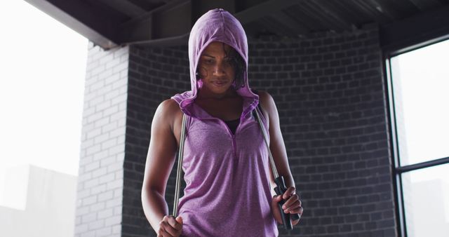 Portrait of african american woman holding skipping rope wearing hoodie looking at camera in an empty urban building. urban fitness and healthy lifestyle.