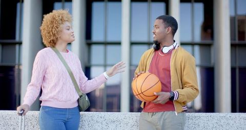 Young diverse friends chatting and leaning at urban plaza holding basketball and suitcase