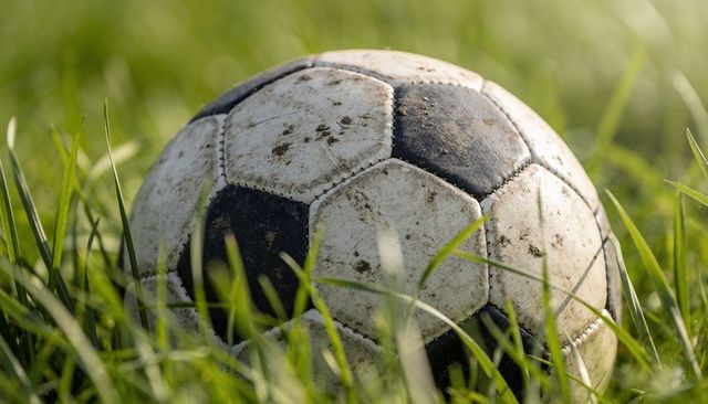 Resting worn soccer ball in sunlit tall grass showing mud scuffs and weathered texture