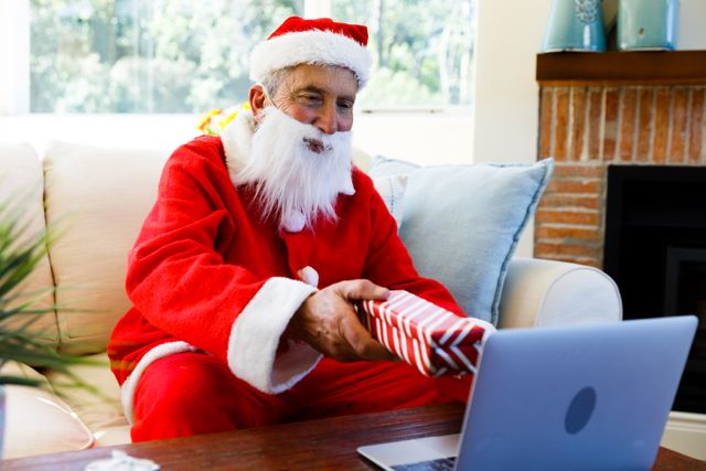Senior man dressed as Santa Claus holding out a Christmas gift during a video call on a laptop. He is sitting on a couch in a cozy living room, spreading holiday cheer. Perfect for illustrating virtual celebrations, online communication during holidays, and festive home settings.