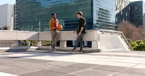 Diverse friends chatting and leaning on barrier holding basketball in sunlit urban plaza