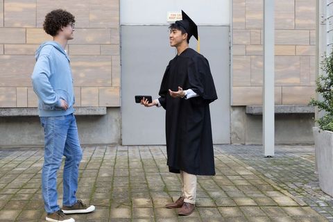 Graduate chatting with friend on campus courtyard holding smartphone and gesturing